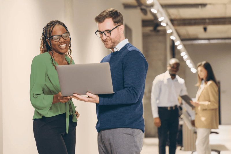 Professional businesspeople collaborating on laptop, coworkers bustling in soft focus hallway workplace environment